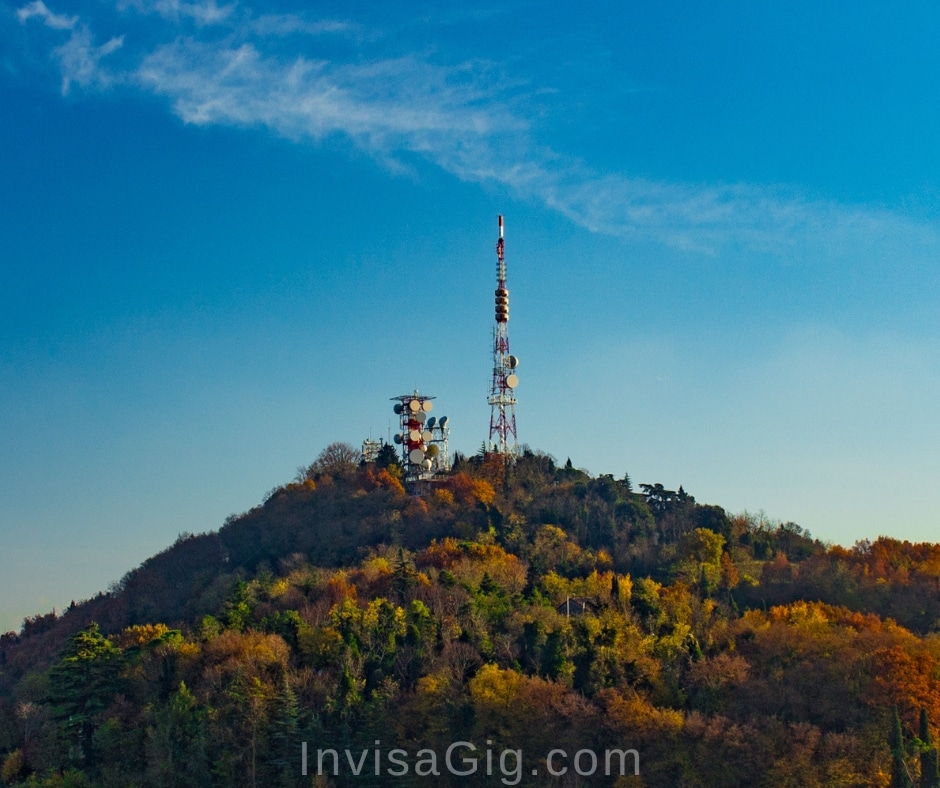 A picture of two cellular towers on a large hilltop covered in trees. Clear blue skies. A very good representation of cellular towers used for getting good cell signal from for products like the InvisaGig.